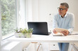 © epiximages - business man with blue shirt and black glasses is standing behind standing table and is working with his tablet and a black laptop in a modern office