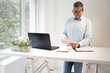 © epiximages - business man with blue shirt and black glasses is standing behind standing table and is working with his tablet and a black laptop in a modern office