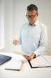 © epiximages - business man with blue shirt and black glasses is standing behind standing table and is working with his tablet and a black laptop in a modern office