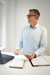 © epiximages - business man with blue shirt and black glasses is standing behind standing table and is working with his tablet and a black laptop in a modern office