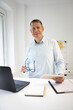© epiximages - business man with blue shirt and black glasses is standing behind standing table and is working with his tablet and a black laptop in a modern office