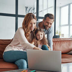 Wall Mural - Young beautiful family with little girl bonding together and smiling while using laptop at home