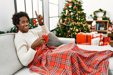 Sticker - Young african american man sitting on the sofa drinking coffee by christmas tree smiling amazed and surprised and pointing up with fingers and raised arms.