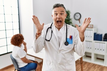 Sticker - Middle age doctor man at the clinic with a patient celebrating crazy and amazed for success with arms raised and open eyes screaming excited. winner concept