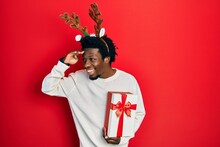 Young African American Man Wearing Deer Christmas Hat Holding Gift Very Happy And Smiling Looking Far Away With Hand Over Head. Searching Concept.