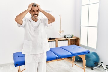 Canvas Print - Middle age hispanic therapist man working at pain recovery clinic suffering from headache desperate and stressed because pain and migraine. hands on head.