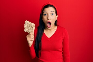 Wall Mural - Young latin woman holding uncooked noodles scared and amazed with open mouth for surprise, disbelief face