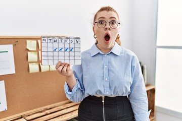 Sticker - Young redhead woman holding travel calendar at the office scared and amazed with open mouth for surprise, disbelief face