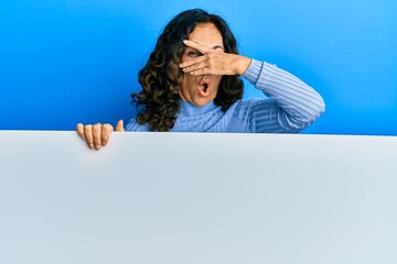 Sticker - Middle age hispanic woman holding blank empty banner peeking in shock covering face and eyes with hand, looking through fingers with embarrassed expression.