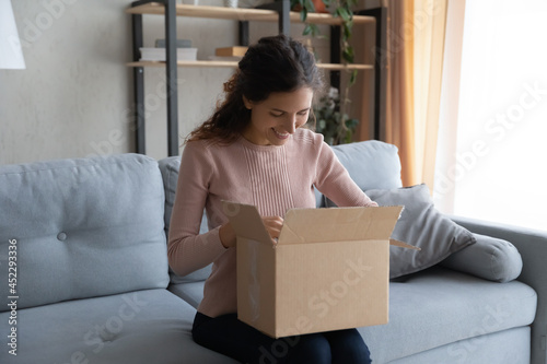 Hispanic woman sit on sofa put on laps box opens parcel feels satisfied with purchased goods on internet, addressee got package from friend. Quick delivery service, trusted transport company concept