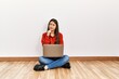 © Krakenimages.com - Young brunette woman sitting on the floor at empty room with laptop serious face thinking about question with hand on chin, thoughtful about confusing idea