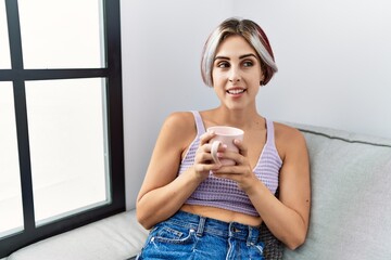 Sticker - Young caucasian girl drinking coffee sitting on the sofa at home.