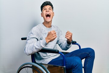 Wall Mural - Young hispanic man sitting on wheelchair angry and mad screaming frustrated and furious, shouting with anger. rage and aggressive concept.