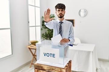 Poster - Hispanic man with beard voting putting envelop in ballot box doing stop sing with palm of the hand. warning expression with negative and serious gesture on the face.