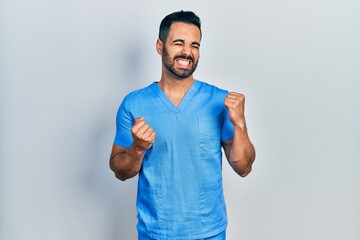 Sticker - Handsome hispanic man with beard wearing blue male nurse uniform excited for success with arms raised and eyes closed celebrating victory smiling. winner concept.