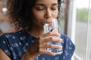 Wall Mural - Peaceful smiling young African American woman drinking pure filtered water with closed eyes, satisfying thirst, enjoying cold beverage, keeping hydration balance, metabolism. Healthy lifestyle