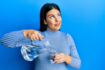 Poster - Beautiful brunette woman pouring water in glass smiling looking to the side and staring away thinking.