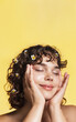 Body positivity and women beauty. Vertical shot of curly girl touches her clean, healthy facial skin with satisfaction, smiling pleased with eyes closed, has chamomile on her hair, yellow background