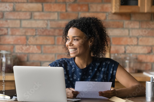Happy millennial Black student girl receiving paper letter, invitation, acceptance notice from college. Young woman holding document from envelope, smiling, looking away, thinking over good news