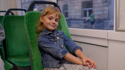 Attractive little girl child kid passenger riding at public modern bus or tram transport, looking out the window. Evening time. City, urban, transportation. Expressions. Positive people. Slow motion