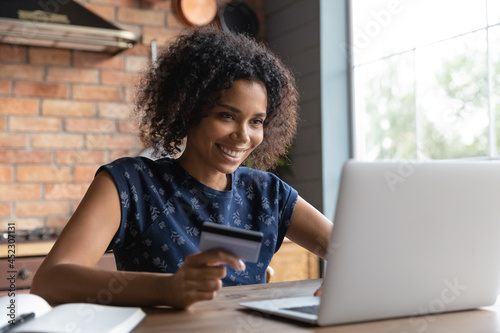 Happy millennial female shopper using laptop and credit card at home. Young African American woman shopping on internet stores, paying bills, loan fees, making payment for online purchases. Ecommerce
