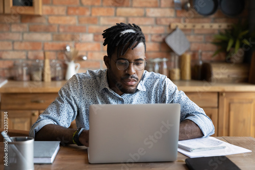 Focused African student guy studying from home, watching learning webinar on laptop, attending virtual class. Serious distance employee, business professional working at computer in kitchen