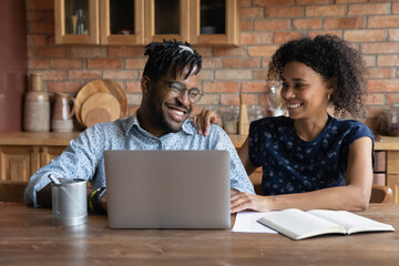 Happy African American couple paying monthly bills, insurance, mortgage fees online on laptop, using computer at home, shopping on internet, getting and discussing good news from online chat