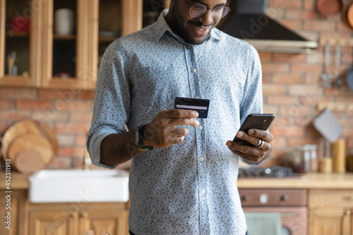 Happy millennial African guy shopping with mobile phone, ordering goods on internet, paying for purchase by credit card, using online banking app for transferring money, getting good cash back