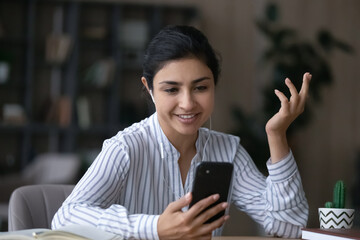 Canvas Print - Smiling young Indian woman in headphones talk speak on video call on cellphone gadget. Happy mixed race female in earphones have webcam virtual digital event on smartphone. Communication concept.