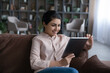 © fizkes - Smiling young Indian woman sit relax on sofa look at tablet screen work online browse internet. Happy millennial ethnic female rest on couch at home talk speak on webcam video call on pad device.
