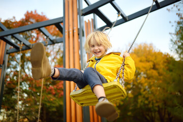 Wall Mural - Little boy having fun on a swing on the playground in public park on autumn day. Happy child enjoy swinging. Active outdoors leisure for child