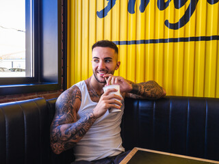 Cheerful attractive Spanish man with tattoos drinking a cocktail with a straw in the restaurant