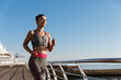 Outdoor shot of young determined fitness woman jogging near the sea, running towards sun along seaside