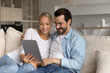 © fizkes - Happy couple with digital tablet making video call from home together, watching or reading online content on internet, getting good news. Smiling husband and wife holding device, looking at screen
