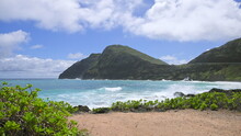 View Of Makapuu Lighthouse. Waves Of Pacific Ocean Wash Over Yellow Sand Of Tropical Beach. Magnificent Mountains Of Hawaiian Island Of Oahu Against Backdrop Of Blue Sky With White Clouds.