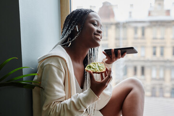 Wall Mural - Side view portrait of young African-American woman recording voice messages while sitting by window and enjoying morning at home, copy space