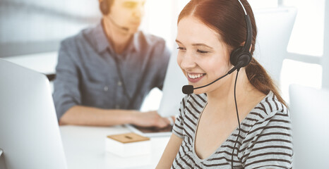 Casual dressed young woman using headset and computer while talking with customers online. Group of operators at work in sunny office. Call center