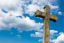 Old Gothic Christian Stone Cross On Dramatic Blue Sky Background With White Clouds. Detail Of Sunlit Ancient Crucifix. Vintage Monument With Moss And Lichen Cover. Idea A Death Or God, Faith And Hope.
