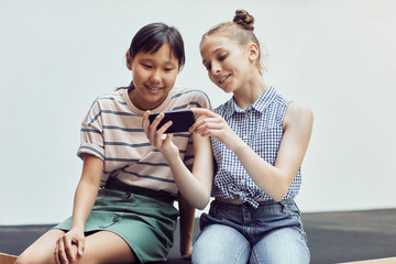 Wall Mural - Minimal portrait of two teenage girls using smartphone and smiling while relaxing against white background