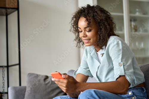 Smiling happy young African American woman holding cell phone sitting on sofa at home, using online mobile apps technology doing online shopping, dating in application, checking social media, texting.