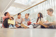 © Seventyfour - Full length portrait of diverse group of children sitting on floor in school and chatting while waiting in hall