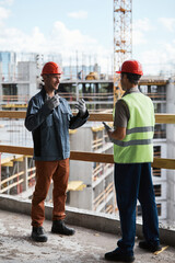 Wall Mural - Vertical full length portrait of two construction workers discussing project while standing at high rise building