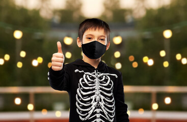 Wall Mural - halloween, holiday and pandemic concept - boy in black protective mask and costume of skeleton showing thumbs up over garland lights at roof top party background