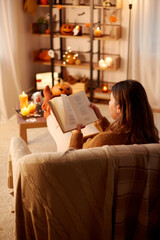 Wall Mural - halloween, holidays and leisure concept - young woman reading book and resting her feet on table at cozy home