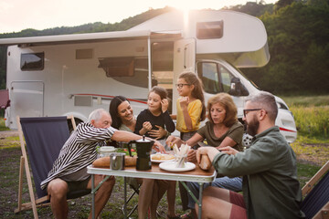 Multi-generation family sitting and eating outdoors by car, caravan holiday trip.