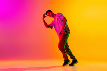 Wall Mural - Full length portrait of young stylish man, break dancing dancer in casual clothes isolated over gradient pink yellow background at dance hall in neon light.