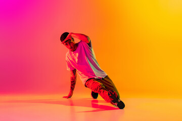 Wall Mural - Portrait of young stylish man, break dancing dancer training in casual clothes isolated over gradient pink yellow background at dance hall in neon light.