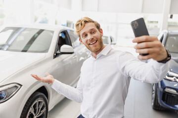 Wall Mural - Man customer male buyer client in white shirt do selfie shot on mobile cell phone show car choose auto want buy new automobile in showroom vehicle dealership store motor show indoor. Sales concept