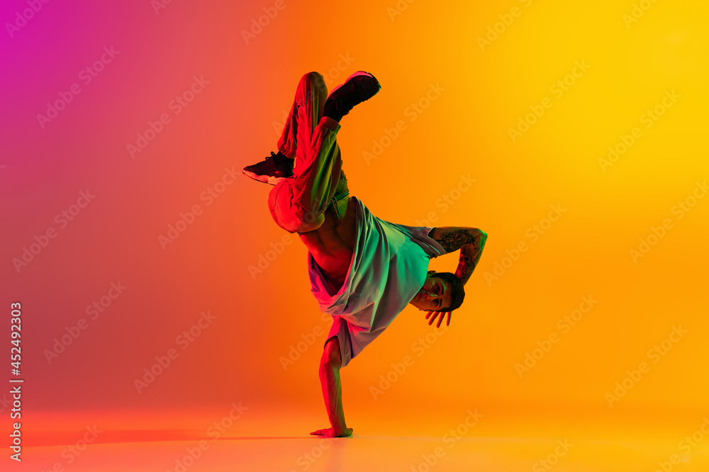 Portrait of young stylish man, break dancing dancer training in casual clothes isolated over gradient pink yellow background at dance hall in neon light. - obrazy, fototapety, plakaty 