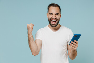 Young overjoyed happy man wear casual basic blank print design white t-shirt using mobile cell phone do winner gesture clench fist isolated on plain pastel light blue color background studio portrait